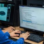 Una persona trabajando en un escritorio con una laptop y un monitor, rodeada de elementos de tecnología y software, en un entorno de oficina moderno y colaborativo.