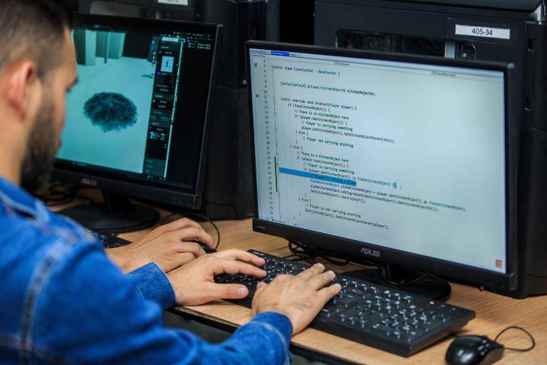 Una persona trabajando en un escritorio con una laptop y un monitor, rodeada de elementos de tecnología y software, en un entorno de oficina moderno y colaborativo.