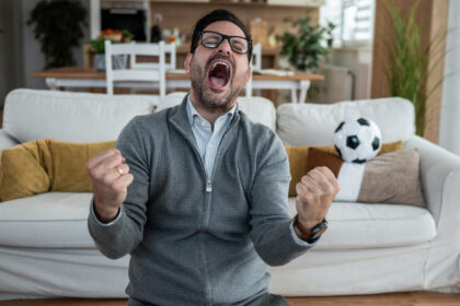 Persona con la mano en el pecho, con una expresión de dolor o alarma, mientras ve un partido de fútbol en la televisión