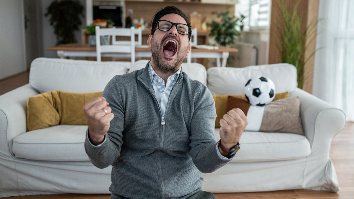 Persona con la mano en el pecho, con una expresión de dolor o alarma, mientras ve un partido de fútbol en la televisión